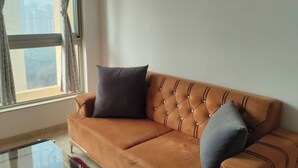 A brown leather sofa with two cushions against a plain wall, a window with curtains, and a clear glass table. - Spacious 1BHK Hiranandani Estate (Thane)