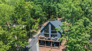 Une maison moderne à deux étages, dotée d’une grande terrasse et d’un espace extérieur couvert, le tout entouré d’une forêt dense.