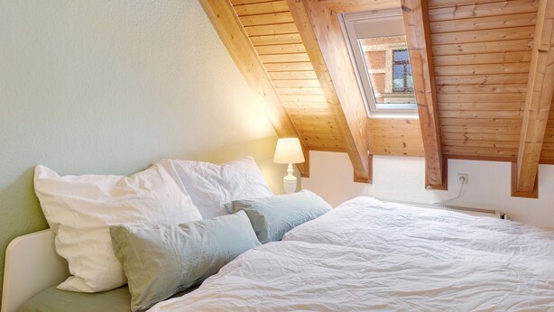 A bedroom with a sloped ceiling, a bed with white bedding, and a nightstand with a lamp.