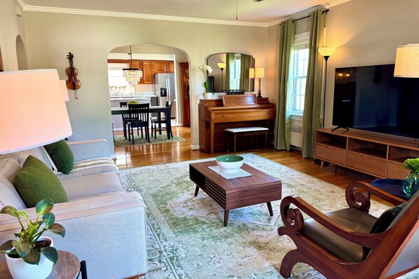 A living room with a sofa, coffee table, and a TV. There is a piano and a dining area in the background. - Westfield Green & Wood Accents & Live Greenery (Richmond)