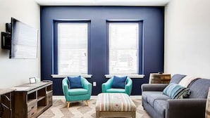 A living room with a dark blue accent wall, two windows, a sofa, a chair, a coffee table, and a TV mounted on the wall.