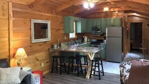 Interior acogedor de una cabaña con techo de madera, cocina con armarios verdes, isla de mármol con taburetes altos y un pequeño comedor.