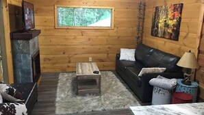 Interior acogedor de una cabaña con chimenea, un sofá de cuero negro, una mesa de centro de madera y una ventana con vistas al bosque.