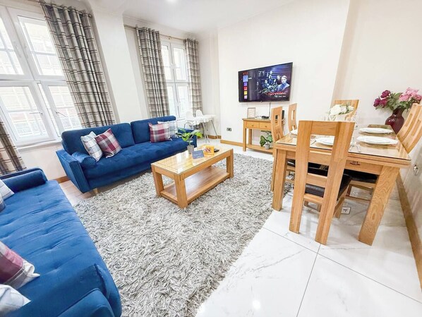 Een woonkamer met een blauwe bank, een salontafel, een eettafel met stoelen en een aan de muur bevestigde televisie.