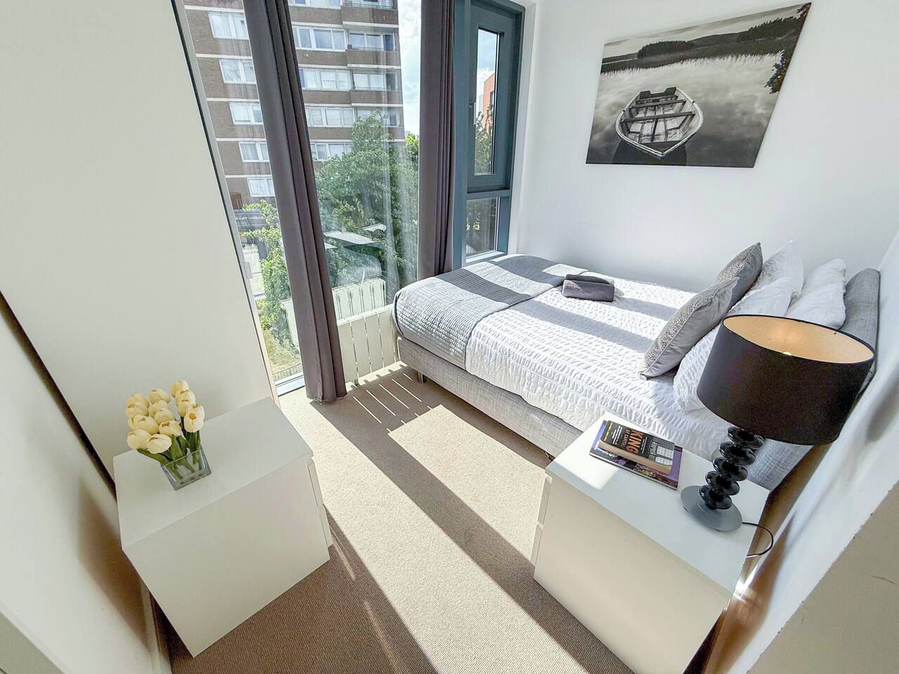 A modern bedroom with a large window, a bed with a striped blanket, a bedside table with a lamp, and a framed picture on the wall.