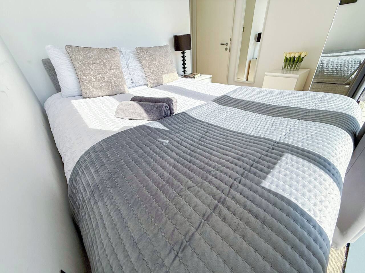 A neatly made bed with a grey quilt and pillows, a bedside table with a lamp, and a mirror reflecting part of the room.