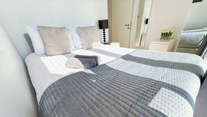 Una cama bien tendida con un edredón gris y almohadas, una mesita de noche con una lámpara, y un espejo que refleja parte de la habitación.