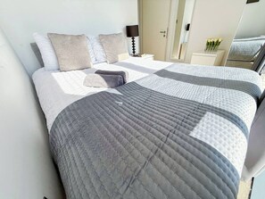 A neatly made bed with a grey quilt and pillows, a bedside table with a lamp, and a mirror reflecting part of the room.