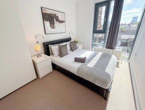 A modern bedroom with a large bed, a nightstand, a bedside lamp, and a view of the city through a large window.