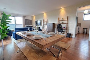 A dining area with a wooden table set for four, a blue sofa, and a TV mounted on the wall. - 3bdr Penthouse W/ Balcony | islington, kings cross (London)
