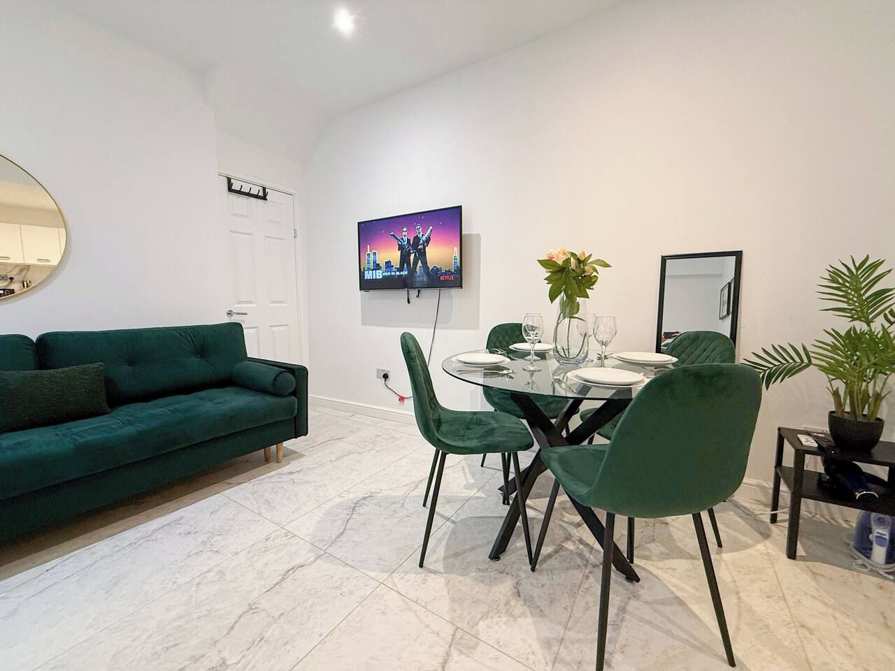 A modern living room with a green sofa, a dining table with chairs, a flat-screen TV mounted on the wall, and a potted plant.