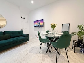 A modern living room with a green sofa, a dining table with chairs, a flat-screen TV mounted on the wall, and a potted plant.