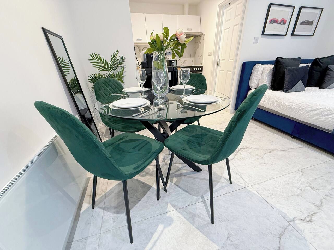 A dining area with a glass table and four green chairs, a vase with flowers, and a kitchen in the background.