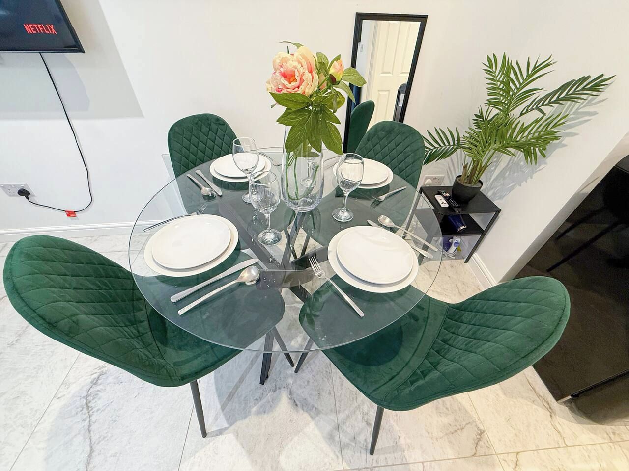 A round glass table with four chairs, a vase with flowers, and a potted plant on a white floor.