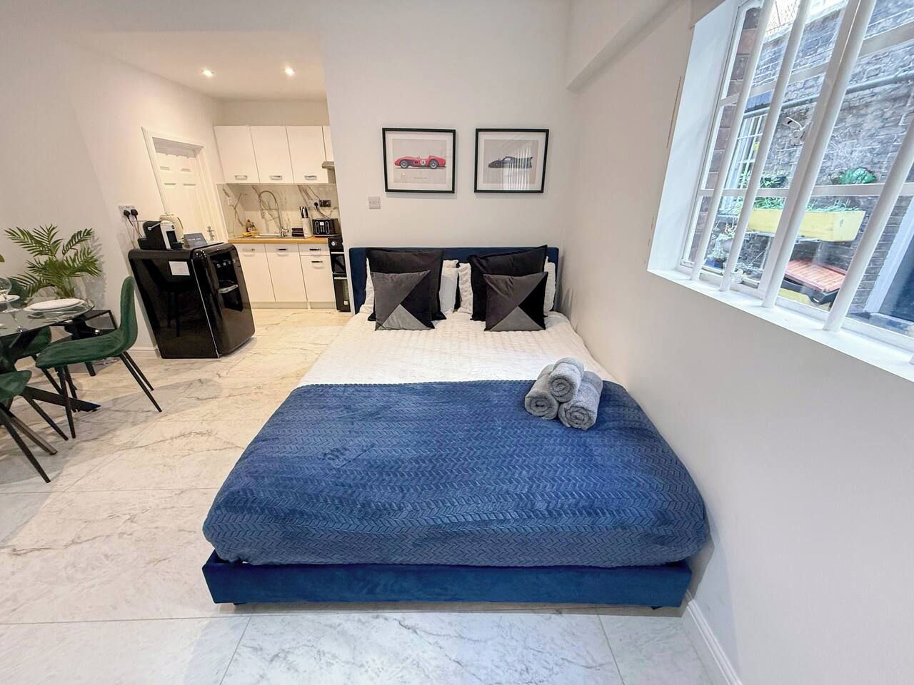 A bedroom with a bed, a window with a view, a kitchen in the background, and a dining area.