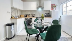 A modern kitchen with white cabinets, a stainless steel trash bin, a dining table with green chairs, and a microwave.
