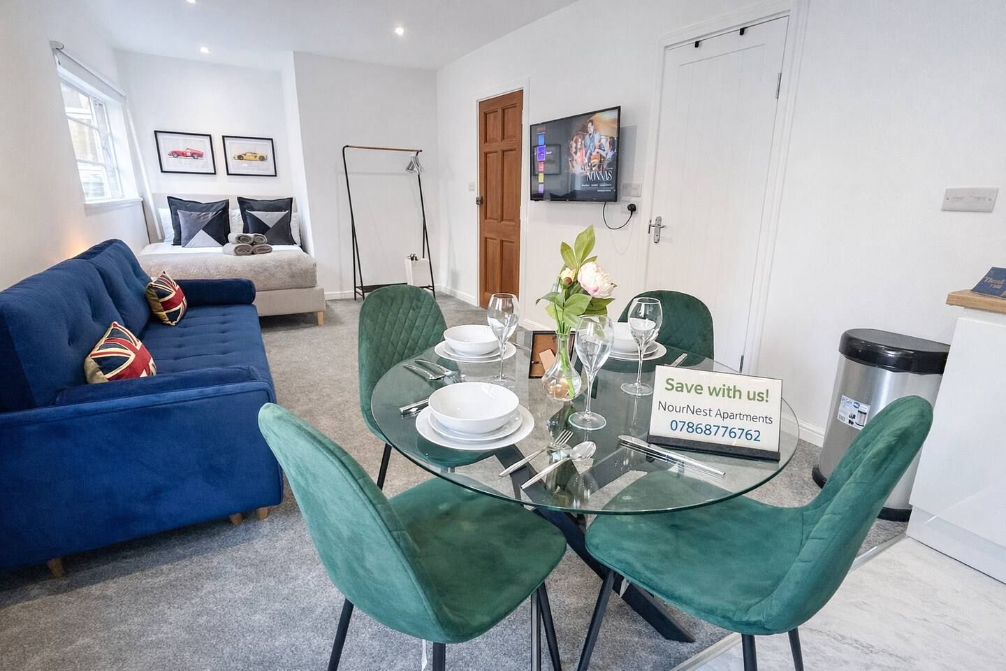 A dining area with a glass table set for four, green upholstered chairs, a sofa with pillows, and a television mounted on the wall.