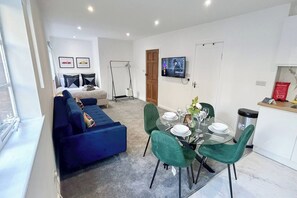A modern living room with a blue sofa, a dining area with a glass table and green chairs, and a TV mounted on the wall.