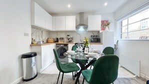 A compact kitchen with white cabinets, a microwave, a toaster, and a sink. There is a dining table with green chairs and a trash bin.