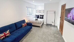 A room with a bed, a blue sofa, a standing fan, a TV, and a window with a city view.
