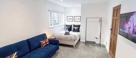 A room with a bed, a blue sofa, a standing fan, a TV, and a window with a city view.
