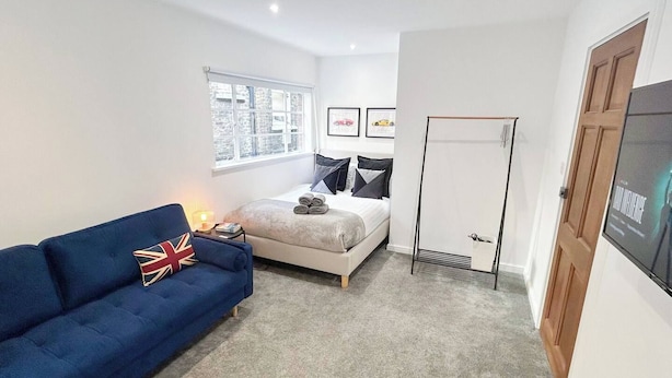 A bedroom with a bed, a blue sofa with a Union Jack pillow, a window with a view of buildings, and a door with a poster.