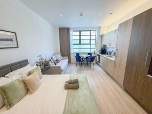 A modern hotel room with a bed, a seating area with a table and chairs, a kitchenette, and a large window. - Chic studio flat in Regent's Park near Marylebone (London)