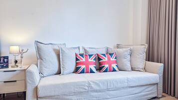 A light grey sofa with three pillows, one with a Union Jack design, in a modern room with a white nightstand, a lamp, and a curtain.