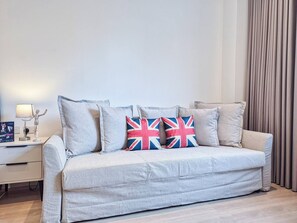 A light grey sofa with three pillows, one with a Union Jack design, in a modern room with a white nightstand, a lamp, and a curtain.