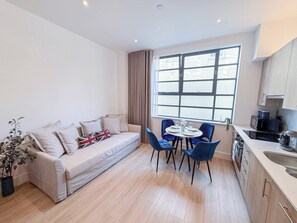 A modern living room with a sofa, a dining table with chairs, a large window, and a kitchen area. - Chic studio flat in Regent's Park near Marylebone (London)