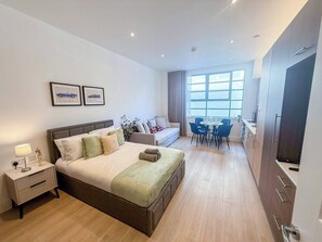 A modern hotel room with a large bed, a bedside table, a desk with chairs, a window with curtains, and a TV.