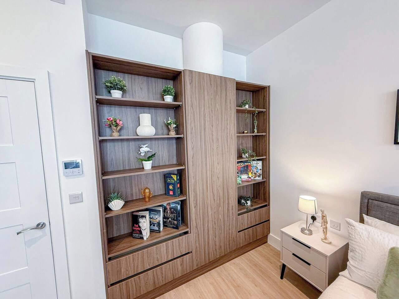 A bedroom with a built-in wooden shelving unit, a bedside table with a lamp, and a bed with pillows.