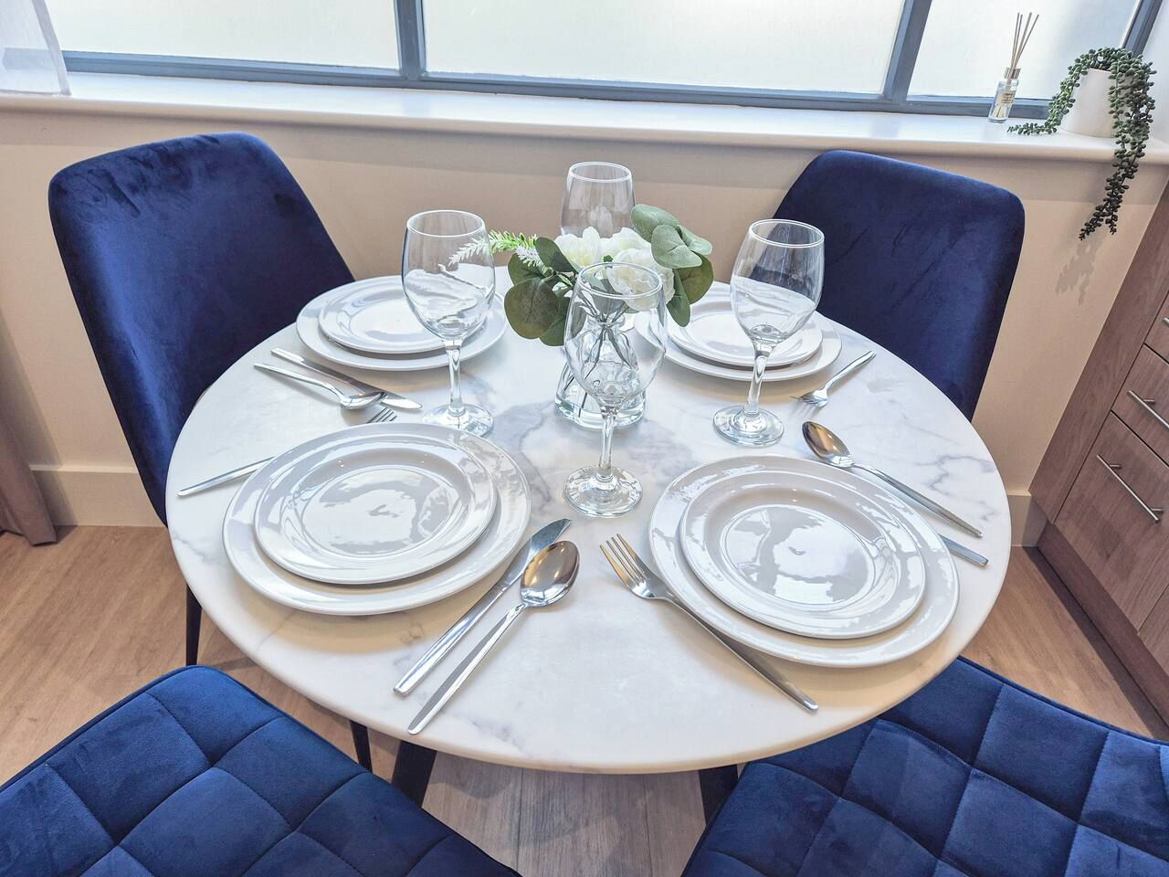 A round dining table set for four with white plates, silverware, and clear glassware.