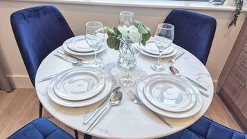 A round dining table set for four with white plates, silverware, and clear glassware.
