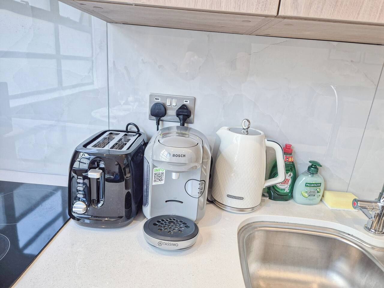 A kitchen with a toaster, coffee maker, kettle, and a sink.