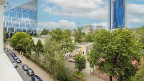 Een modern stedelijk gebied met een combinatie van gebouwen, waaronder een hoge blauwe glazen wolkenkrabber en een lager gebouw in beige tint. Er staan bomen en geparkeerde auto's langs de straat.