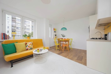A bright living room with a yellow couch, white flooring, and a small round dining table in the corner. The space feels cheerful and modern.