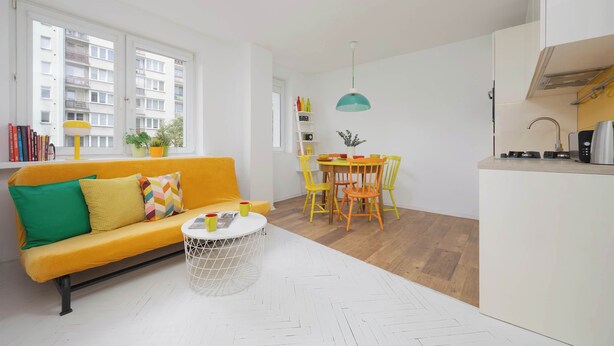 Een lichte woonkamer met een gele bank, een witte salontafel en een eethoek met gele stoelen.