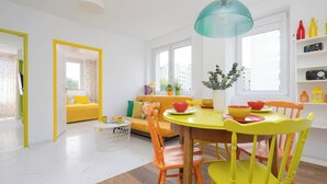 Een lichte, moderne leefruimte met een gele eettafel, oranje en gele stoelen, een gele bank en een groot raam met uitzicht op gebouwen.