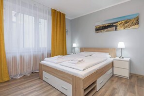 Ein ordentlich bezogenes Bett mit zwei Kissen und gefalteten Handtüchern, ein Nachttisch mit Lampe und ein Bild an der Wand.