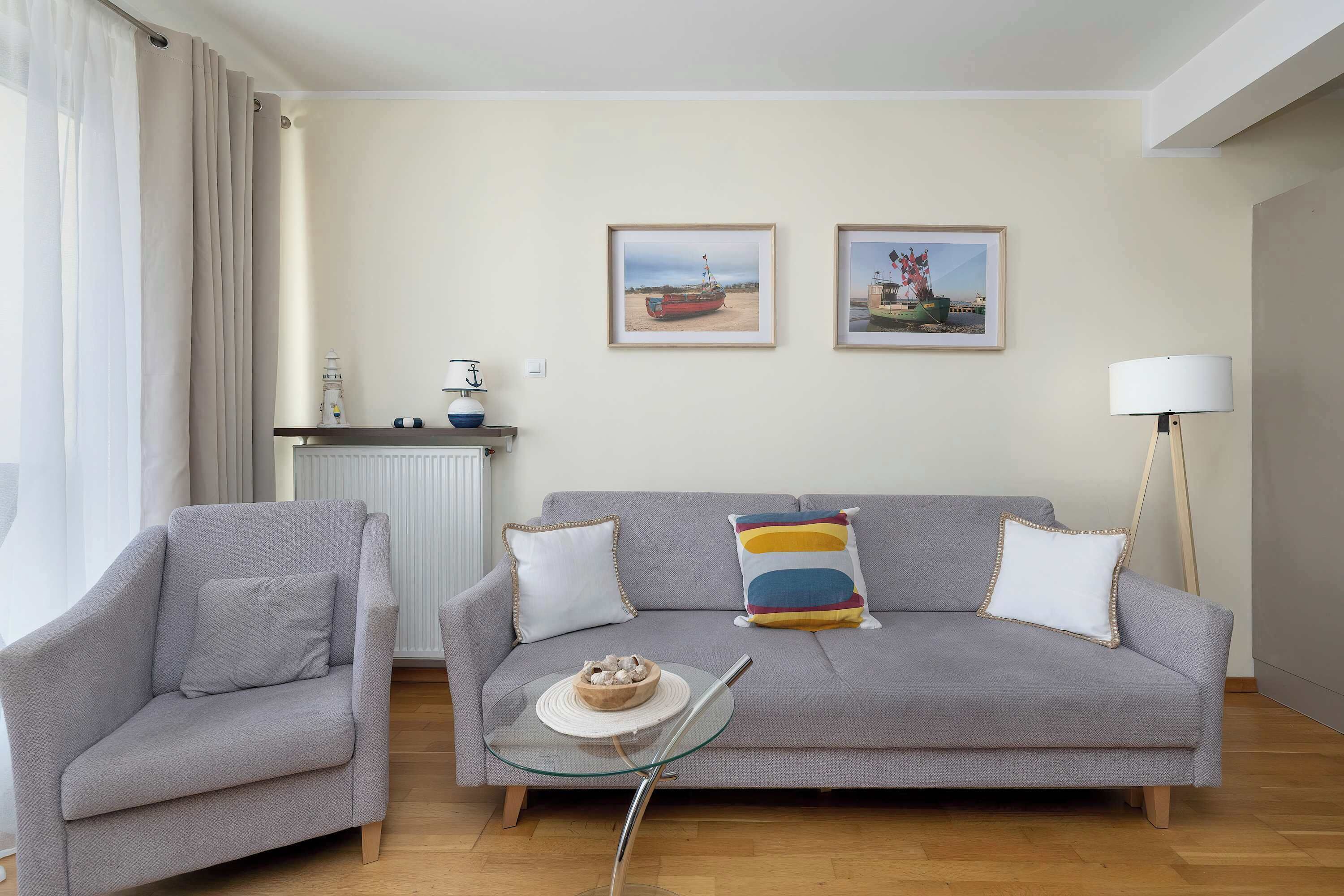 A living room with a grey sofa, a grey armchair, a glass coffee table, and two framed pictures on the wall.