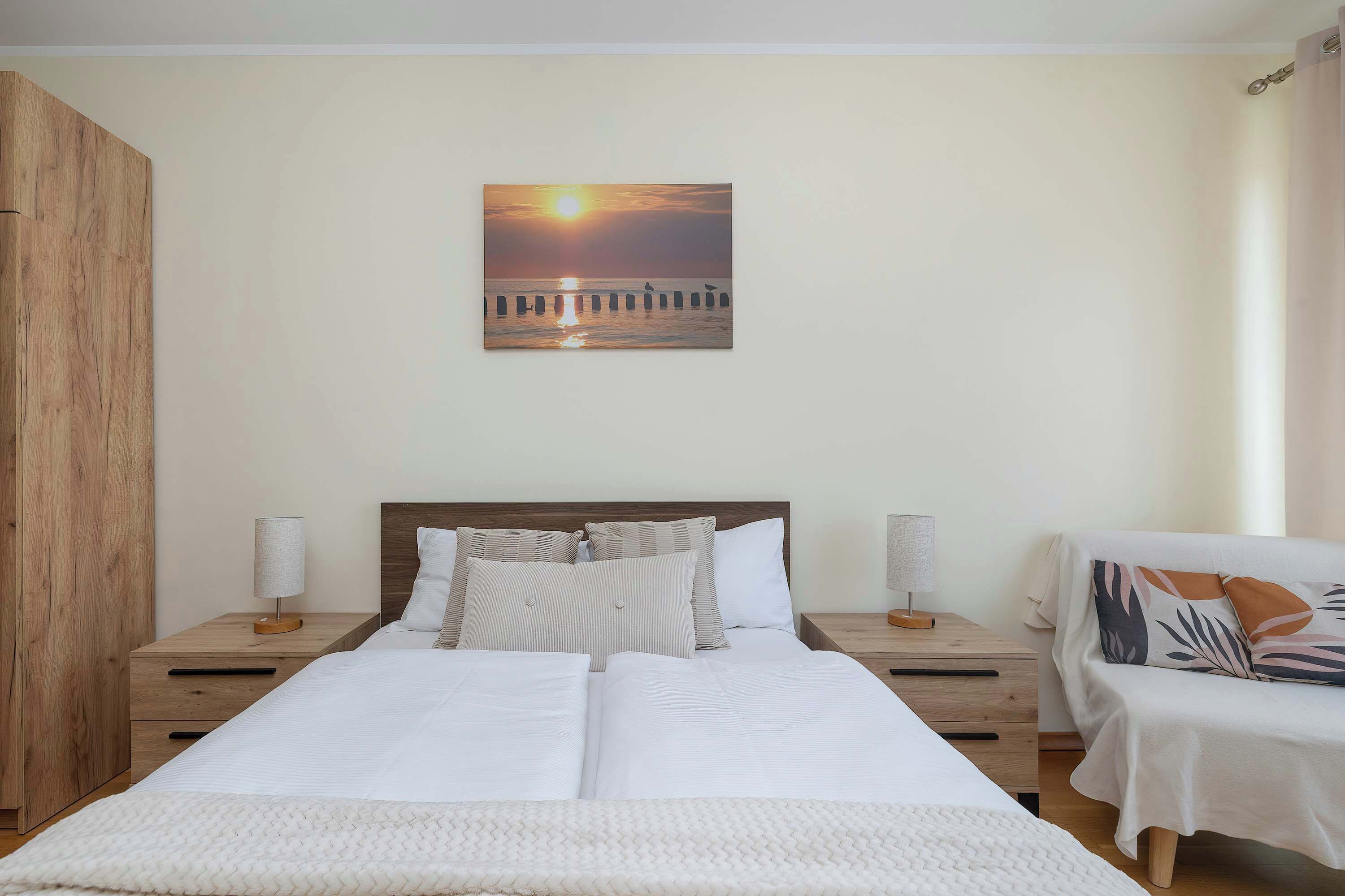 Une chambre avec un grand lit, deux tables de chevet, une armoire en bois et un tableau représentant un coucher de soleil sur une jetée, accroché au mur.