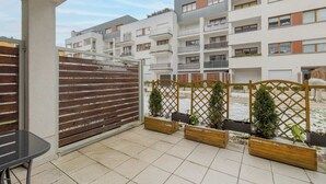 Ein Balkon mit Blick auf Wohnhäuser, Holz-Pflanzkästen mit kleinen Bäumen und einem Metalltisch.
