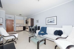 A modern living room with a glass coffee table, white sofas, and a dining area with a kitchen in the background.