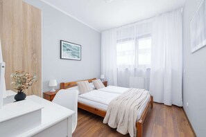 Une chambre à coucher comprenant un lit, des tables de chevet, un bureau, une chaise, une fenêtre avec des rideaux et un tableau encadré accroché au mur.