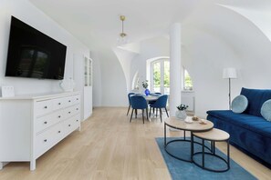A modern living room with a flat-screen TV, a blue sofa, a dining area with chairs, and a large window.