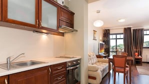 A modern kitchen with wooden cabinets, a built-in oven, and a sink. There is a dining area with a sofa and chairs, and a TV mounted on the wall.