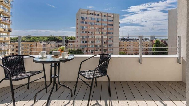 A balcony with two chairs and a small table, overlooking a cityscape with buildings and a clear sky.