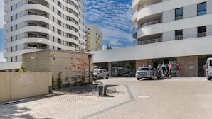A modern building with a curved facade, a paved courtyard, and a parking area with cars.