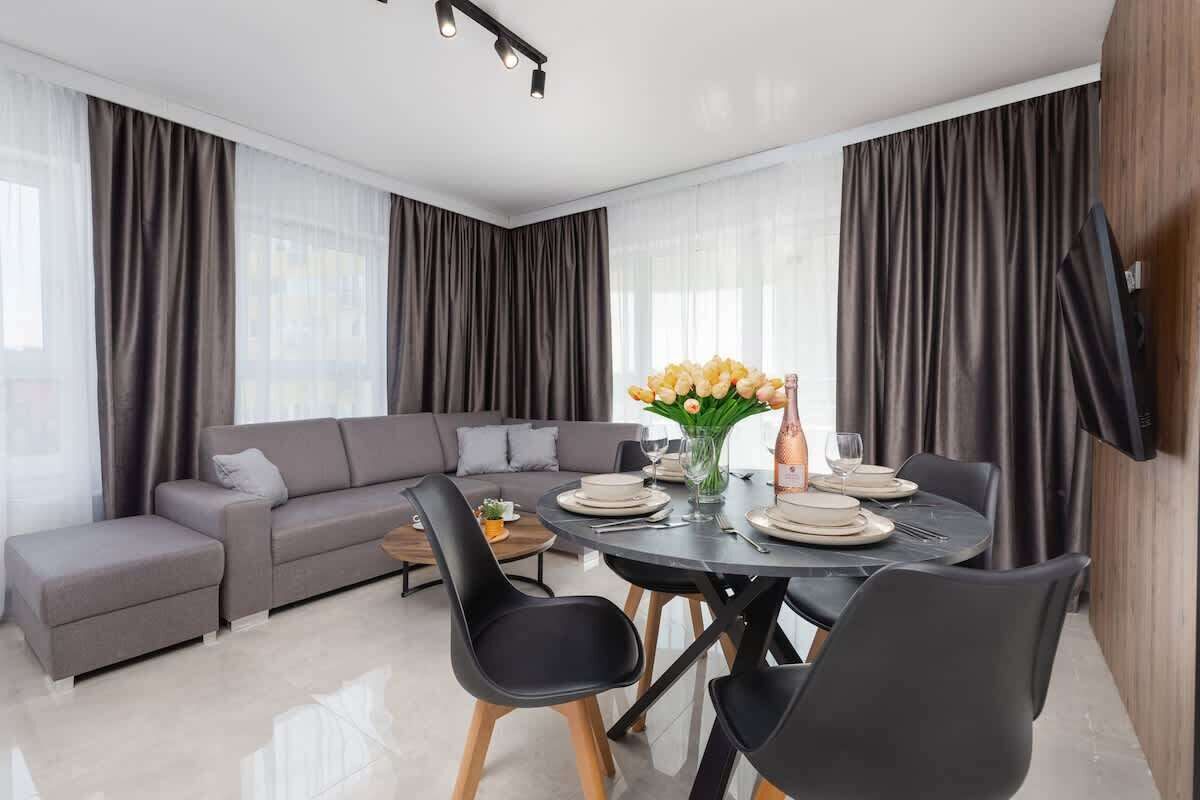 Un salón moderno con un sofá gris, sillas negras, una mesa de comedor para cuatro personas y un jarrón con flores.